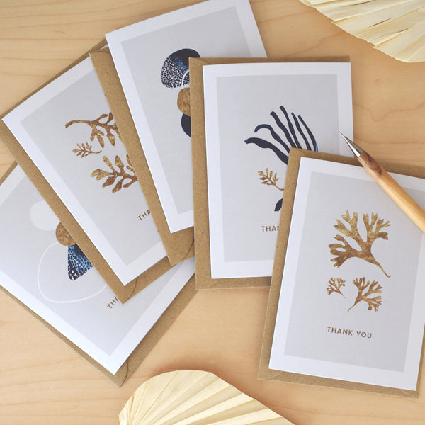 A beautiful set of 5 Thank You note cards, complete with kraft envelopes. Card designs feature blue and gold beach inspired illustrations, including seaweed and pebbles.